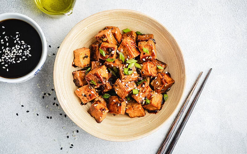 In einer Schale ist als Fleischersatz gebratener marinierter Tofu in Würfelform mit Sojasauce und Frühlingszwiebeln angerichtet.