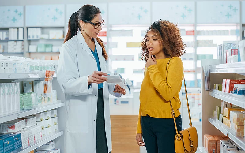 Eine Frau in einem gelben Oberteil informiert sich bei einer Apothekerin über ein Präparat.