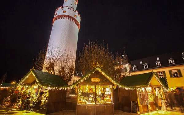 Drei beleuchtete Weihnachtsmarktstände, dahinter weißer Turm mit rotem Stuck, nächtliche Altstadt.