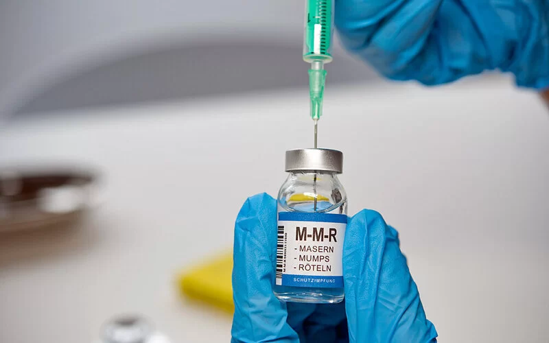 Zwei Hände in Schutzhandschuhen ziehen eine Spritze mit Impfserum gegen Masern-Mumps-Röteln auf.