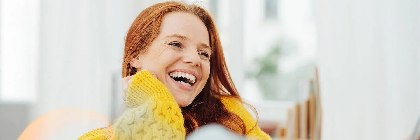 Eine junge, erwachsene Frau mit langen roten Haaren und einem gelben Strickpullover lacht herzhaft mit offenem Mund. Sie hat sehr schöne, weiße Zähne.