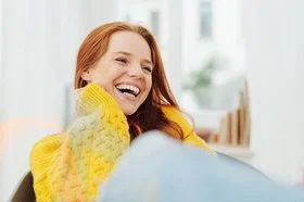 Eine junge, erwachsene Frau mit langen roten Haaren und einem gelben Strickpullover lacht herzhaft mit offenem Mund. Sie hat sehr schöne, weiße Zähne.