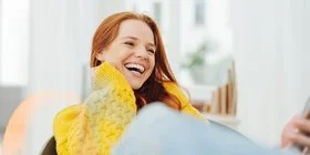 Eine junge, erwachsene Frau mit langen roten Haaren und einem gelben Strickpullover lacht herzhaft mit offenem Mund. Sie hat sehr schöne, weiße Zähne.