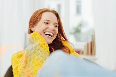 Eine junge, erwachsene Frau mit langen roten Haaren und einem gelben Strickpullover lacht herzhaft mit offenem Mund. Sie hat sehr schöne, weiße Zähne.