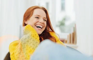 Eine junge, erwachsene Frau mit langen roten Haaren und einem gelben Strickpullover lacht herzhaft mit offenem Mund. Sie hat sehr schöne, weiße Zähne.