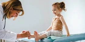 Eine Kinderärztin untersucht die Füße eines etwa 3-jährigen Mädchens, das auf einer Untersuchungsliege sitzt.