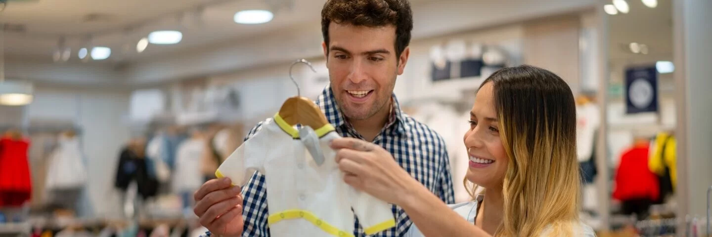 Ein Mann und eine schwangeren Frau schauen sich in einem großen Kinderkaufhaus Babykleidung für die Erstausstattung an.