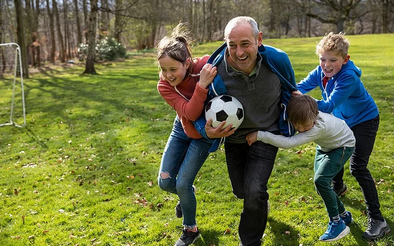 Ein älterer Mann läuft mit einen Fußball in der Hand, drei Kinder halten sich an ihm fest.