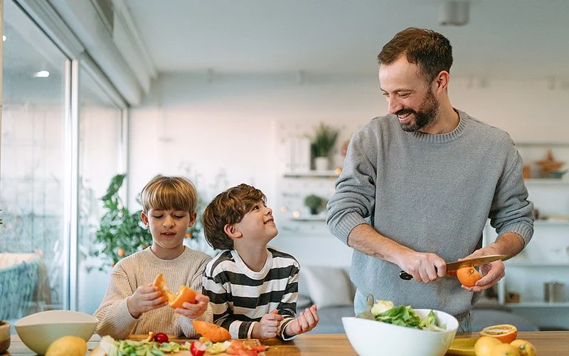 Ein Vater steht mit seinen beiden Söhnen in der Küche und bereitet eine gesunde Mahlzeit mit frischem Obst und Gemüse vor.
