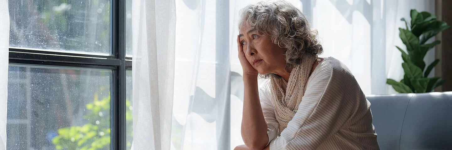 Eine ältere Frau sitzt auf der Couch und schaut aus dem Fenster, mit einer Hand stützt sie ihren Kopf ab.