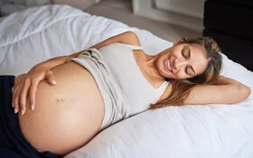 Schwangere liegt auf dem Bett und massiert mit einer Hand sanft ihren Bauch.