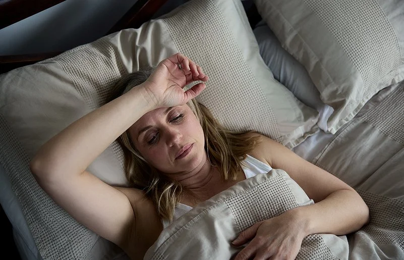 Eine Frau liegt mit geöffneten Augen im Bett und hält sich den Arm über die Stirn.