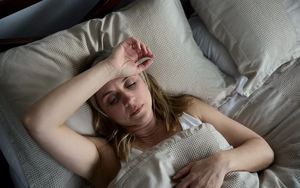 Eine Frau liegt mit geöffneten Augen im Bett und hält sich den Arm über die Stirn.
