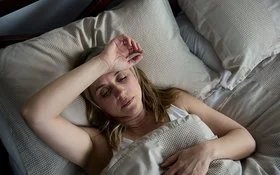 Eine Frau liegt mit geöffneten Augen im Bett und hält sich den Arm über die Stirn.