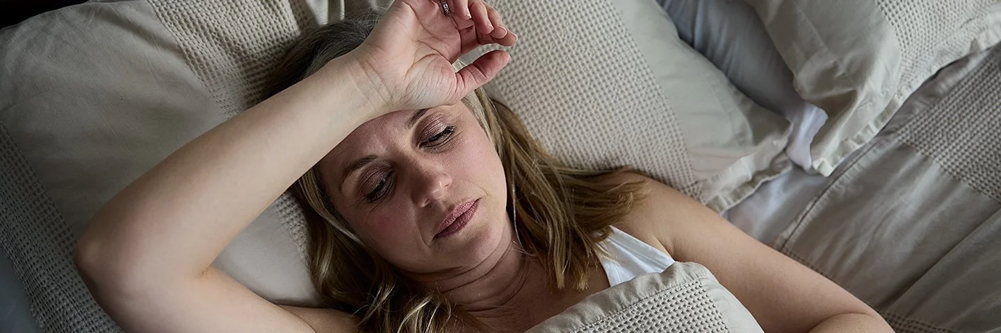 Eine Frau liegt mit geöffneten Augen im Bett und hält sich den Arm über die Stirn.