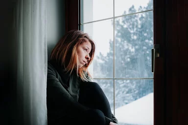 Eine junge Frau sitzt am Fenster und schaut nach draußen in eine verregnete Landschaft. Ihr Blick ist niedergeschlagen.