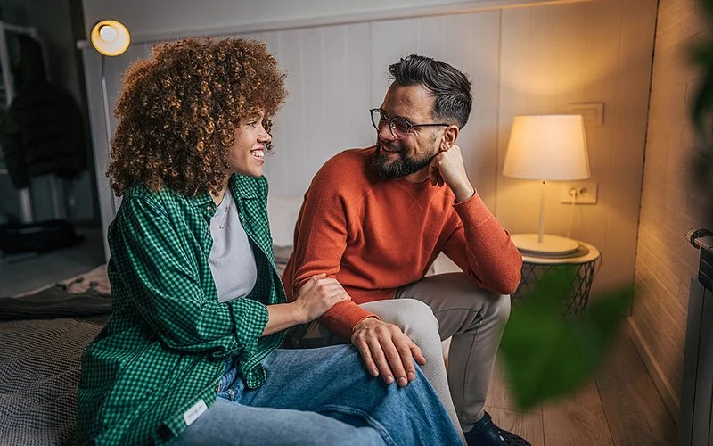 Zwei Personen sitzen auf einem Bett, die Frau trägt ein kariertes Hemd und Jeans, der Mann einen orangenen Pullover und Brille, sie lächeln sich an.