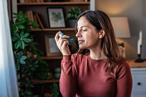 Eine Frau mit langem Haar und rotem Pullover hält ein Inhalationsgerät in der Hand, im Hintergrund sind ein Bücherregal, mehrere Zimmerpflanzen und eine Lampe zu erkennen.