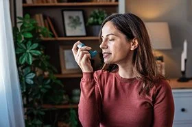 Eine Frau mit langem Haar und rotem Pullover hält ein Inhalationsgerät in der Hand, im Hintergrund sind ein Bücherregal, mehrere Zimmerpflanzen und eine Lampe zu erkennen.