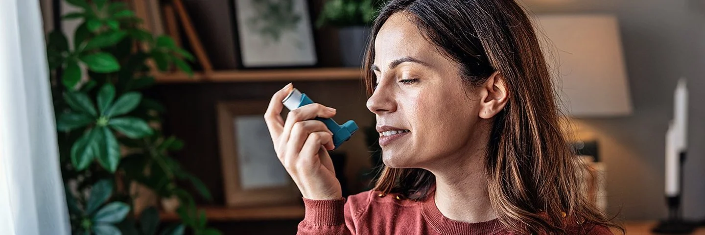 Eine Frau mit langem Haar und rotem Pullover hält ein Inhalationsgerät in der Hand, im Hintergrund sind ein Bücherregal, mehrere Zimmerpflanzen und eine Lampe zu erkennen.
