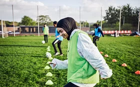 Eine Frau mit Kopftuch und grüner Weste treibt Sport auf einem Fußballfeld.