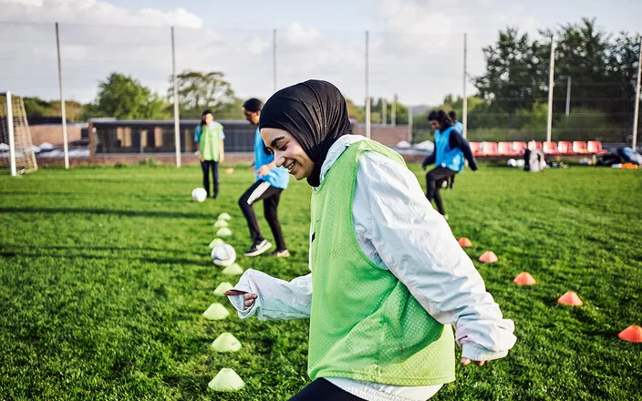 Eine Frau mit Kopftuch und grüner Weste treibt Sport auf einem Fußballfeld.