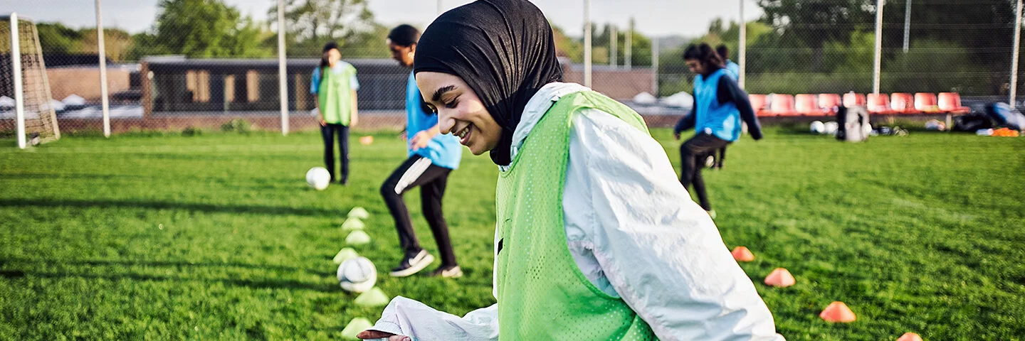 Eine Frau mit Kopftuch und grüner Weste treibt Sport auf einem Fußballfeld.