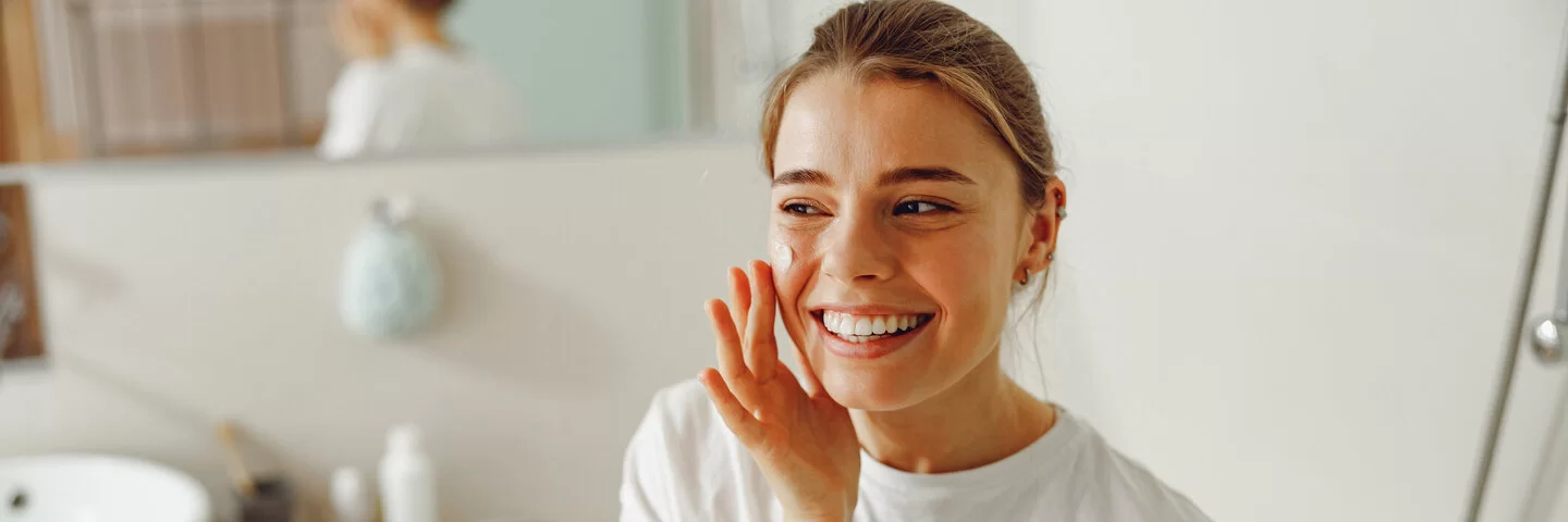 Eine junge Frau steht im Badezimmer und cremt sich lächelnd ihr Gesicht ein.