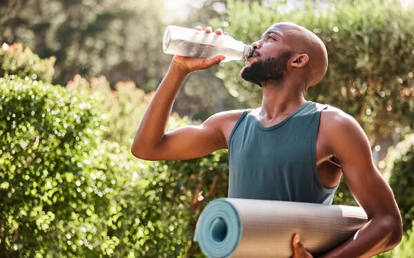 Ein junger Mann trinkt nach dem Sport im Freien aus einer Glasflasche Wasser.