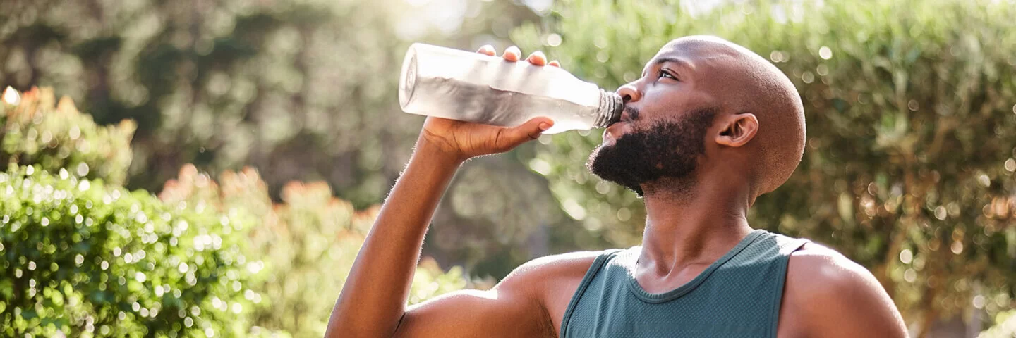 Ein junger Mann trinkt nach dem Sport im Freien aus einer Glasflasche Wasser.