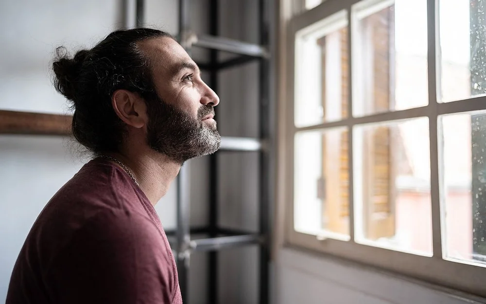 Ein Mann befindet sich in einem Innenraum und schaut aus einem Fenster. Er lächelt.
