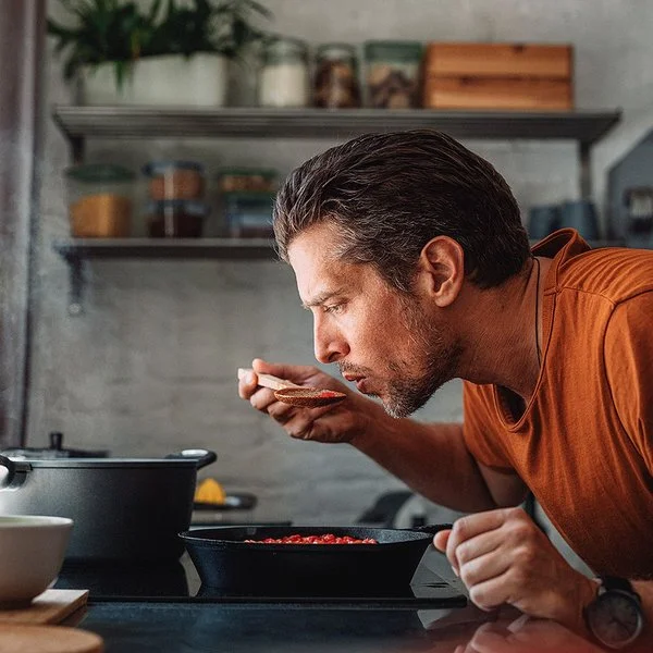 Ein Mann mit orangenem T-Shirt steht in der Küche und kocht. Er beugt sich über den Topf und probiert mit einem Löffel sein Essen.