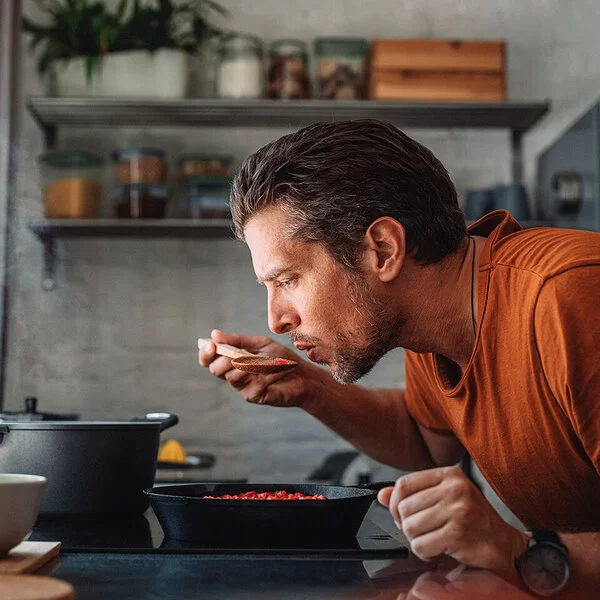Ein Mann mit orangenem T-Shirt steht in der Küche und kocht. Er beugt sich über den Topf und probiert mit einem Löffel sein Essen.