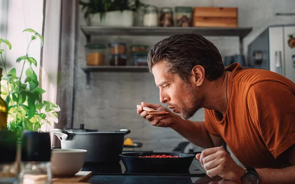 Ein Mann mit orangenem T-Shirt steht in der Küche und kocht. Er beugt sich über den Topf und probiert mit einem Löffel sein Essen.