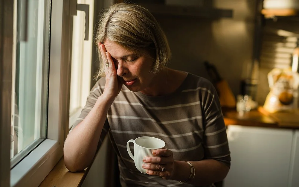 Eine Frau steht am Fenster eines Wohnzimmers. Ihr rechter Ellenbogen ist auf der Fensterbank abgestützt, während sie den Kopf in die rechte Hand stützt. Ihre Augen sind geschlossen. In der linken Hand hält sie eine Tasse.