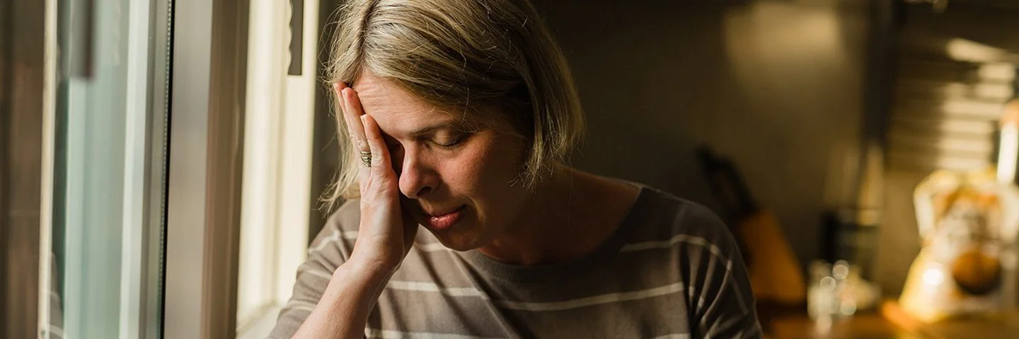 Eine Frau steht am Fenster eines Wohnzimmers. Ihr rechter Ellenbogen ist auf der Fensterbank abgestützt, während sie den Kopf in die rechte Hand stützt. Ihre Augen sind geschlossen. In der linken Hand hält sie eine Tasse.