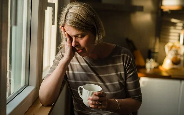 Eine Frau steht am Fenster eines Wohnzimmers. Ihr rechter Ellenbogen ist auf der Fensterbank abgestützt, während sie den Kopf in die rechte Hand stützt. Ihre Augen sind geschlossen. In der linken Hand hält sie eine Tasse.