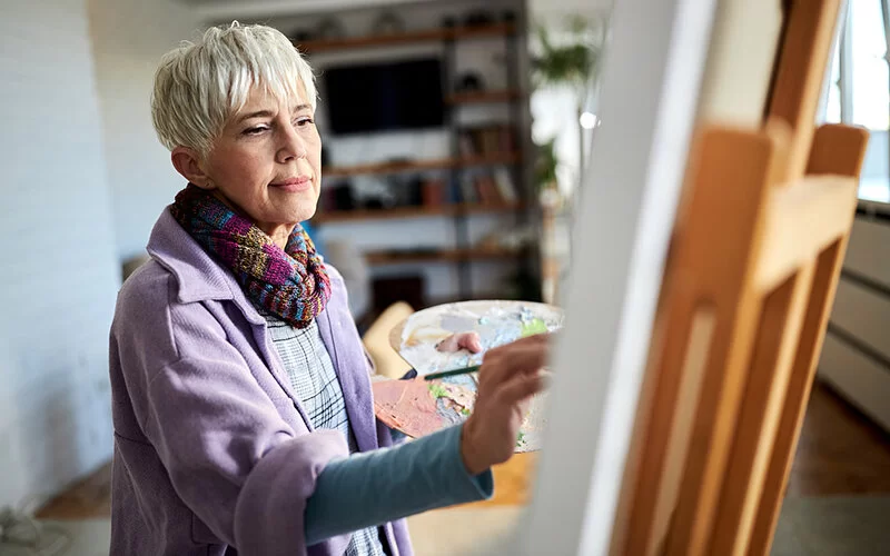 Eine ältere Frau malt ein Bild an einer Staffelei.