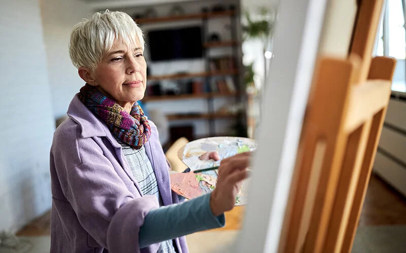 Eine ältere Frau malt ein Bild an einer Staffelei.