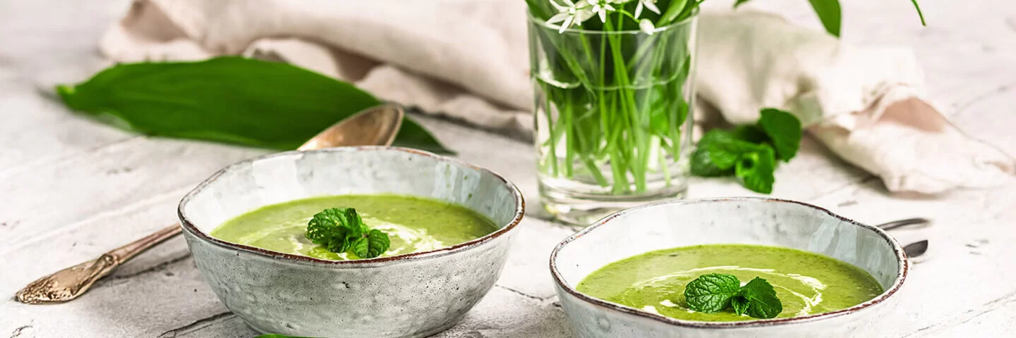 Zwei Keramikschalen mit cremiger Bärlauchsuppe. Daneben ein Strauß aus Bärlauchblättern mit weißen Blüten auf einem hellen, rustikalen Tisch.