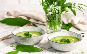 Zwei Keramikschalen mit cremiger Bärlauchsuppe. Daneben ein Strauß aus Bärlauchblättern mit weißen Blüten auf einem hellen, rustikalen Tisch.