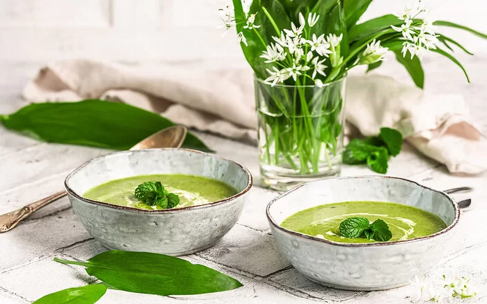Zwei Keramikschalen mit cremiger Bärlauchsuppe. Daneben ein Strauß aus Bärlauchblättern mit weißen Blüten auf einem hellen, rustikalen Tisch.