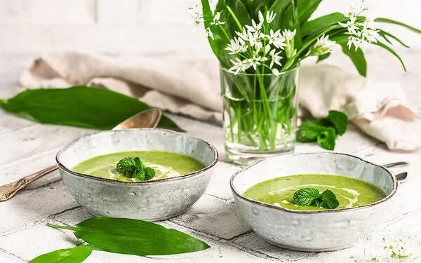 Zwei Keramikschalen mit cremiger Bärlauchsuppe. Daneben ein Strauß aus Bärlauchblättern mit weißen Blüten auf einem hellen, rustikalen Tisch.