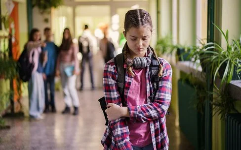 Ein Mädchen im Teenager-Alter steht mit gesenktem Blick auf einem Schulflur.