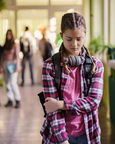 Ein Mädchen im Teenager-Alter steht mit gesenktem Blick auf einem Schulflur.