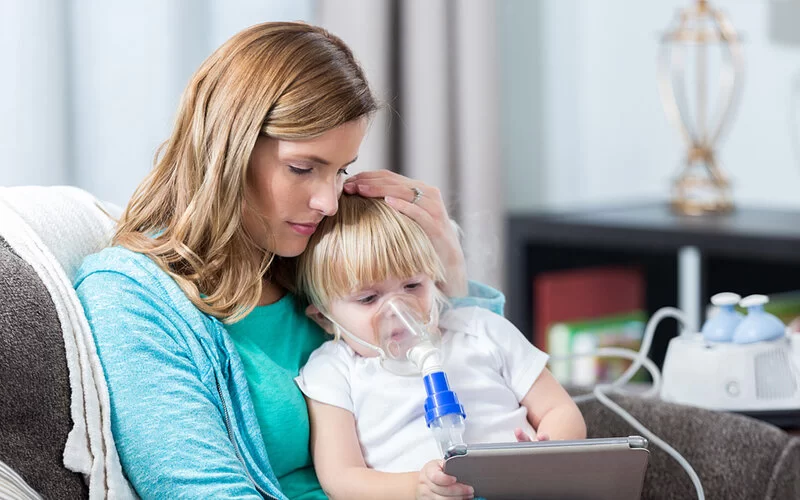 Ein Kind mit der Erbkrankheit Mukoviszidose inhaliert mit einem speziellen Inhalator und sitzt dabei auf dem Schoß seiner Mutter.