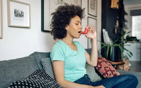 Afroamerikanische, junge Frau sitzt zuhause auf dem Sofa und nutzt ein Asthmaspray mit Kortison.