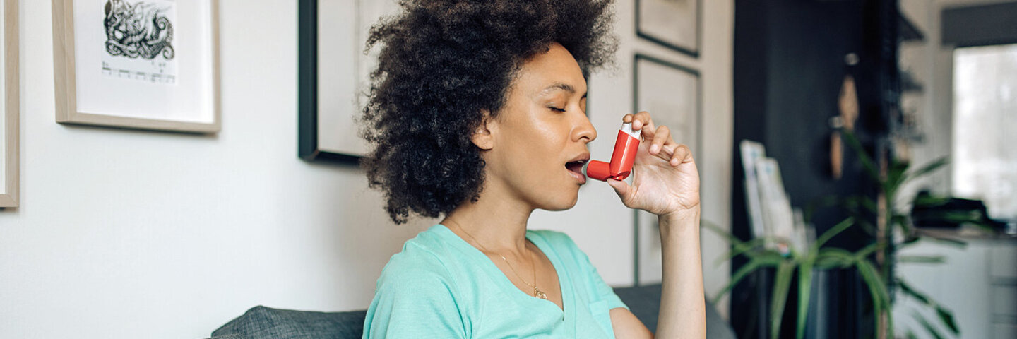 Afroamerikanische, junge Frau sitzt zuhause auf dem Sofa und nutzt ein Asthmaspray mit Kortison.