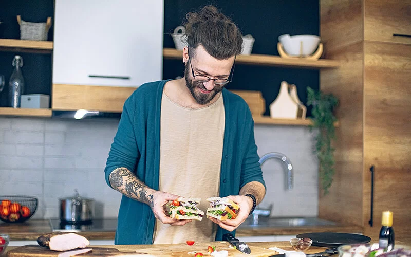 Ein jüngerer Mann mit Tätowierungen am Unterarm und langen braunen Haaren, die zu einem Knoten hochgebunden sind, hält lächelnd in jeder Hand eine Hälfte eines Sandwichs, das er gerade mit vielen frischen Zutaten zubereitet hat.