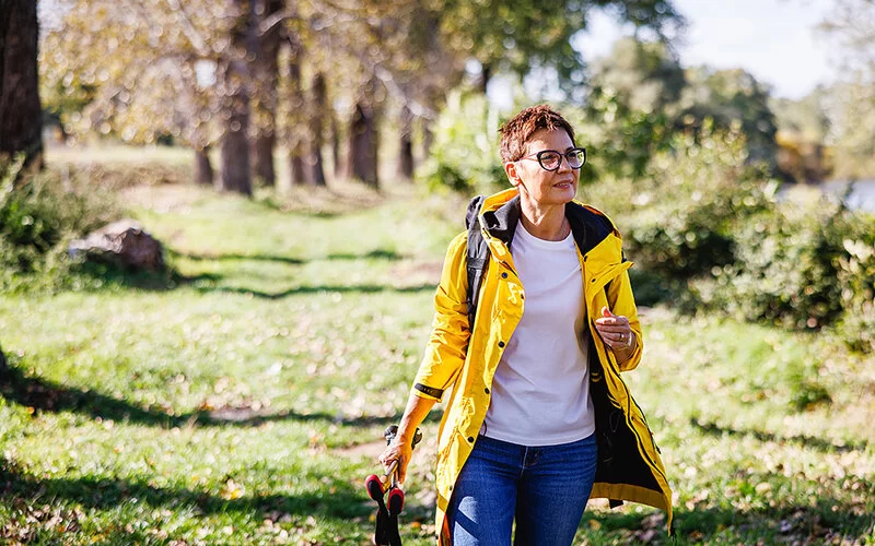 Eine Frau mittleren Alters spaziert mit Nordic-Walking-Stöcken in der Hand bei Sonnenschein durch einen Wald.